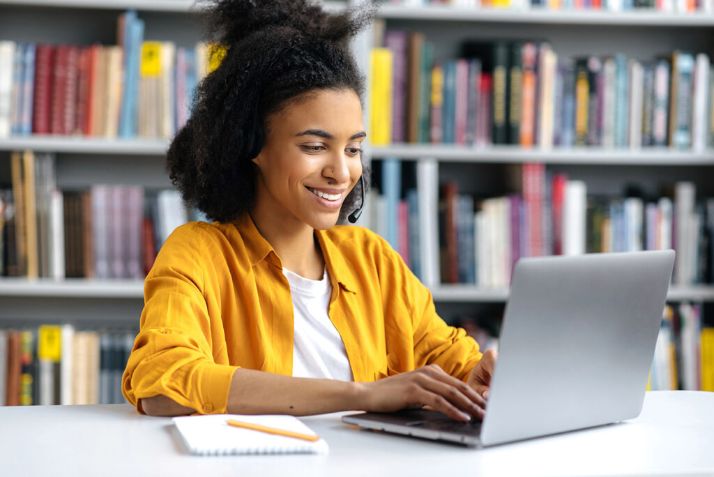 Happy attractive african american girl in stylish clothes, female student sits in the university library at the desk, preparing for the exam or lesson, browsing internet, searching information, smiles